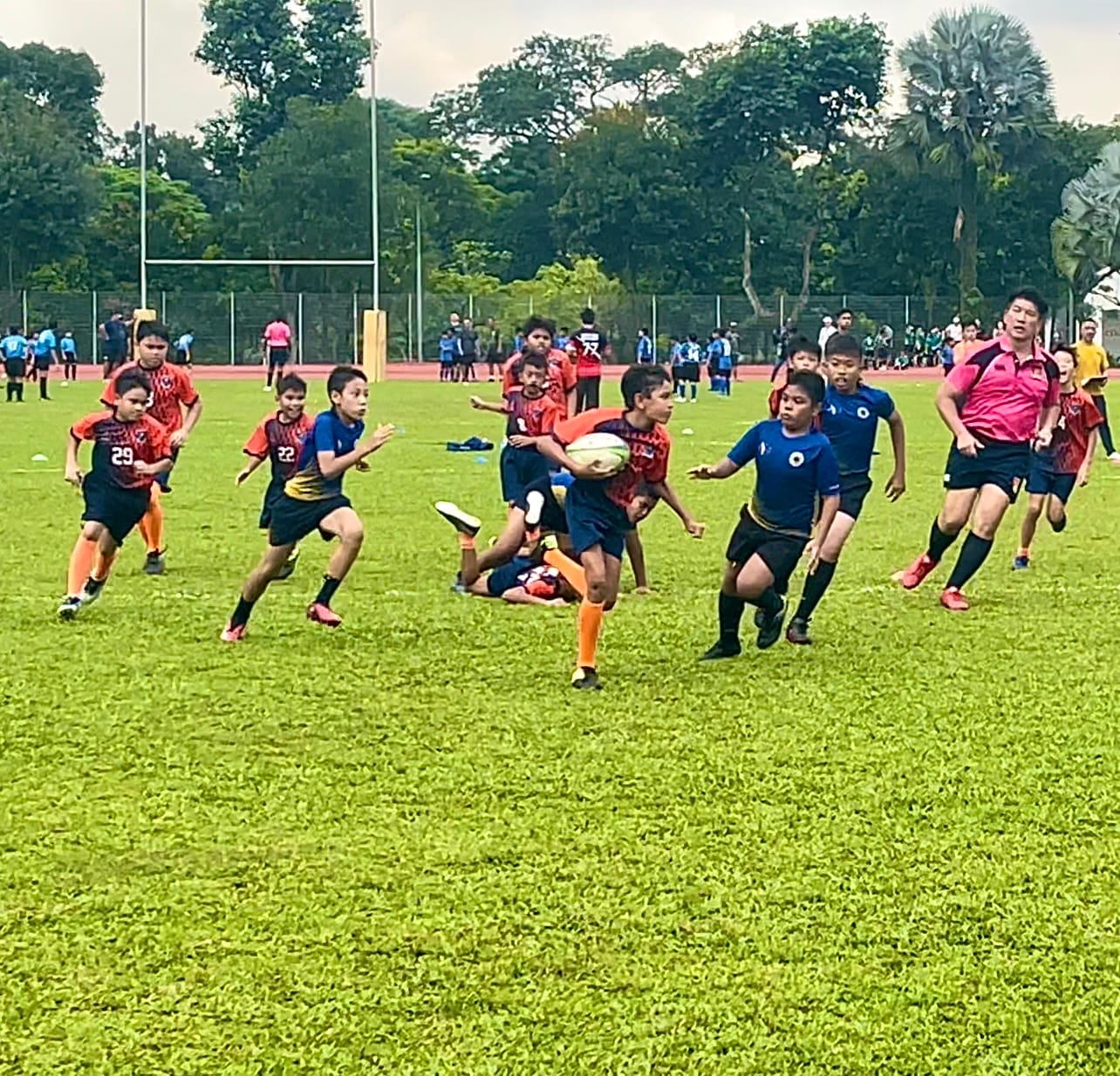 Young boys in red and blue jerseys play rugby on a grass field, with one child carrying the ball while others chase.